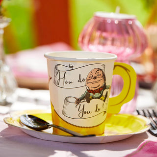 A white mug with a yellow handle and saucer, part of the Alice in Wonderland Bright Tea Cup Set by Talking Tables, featuring an illustration and the text "How do You do?" on a table with a spoon, against a blurred green background.