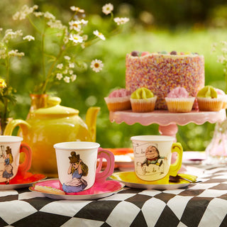 A vibrant tea party setup featuring the Talking Tables "Alice in Wonderland Bright Tea Cup Set," including illustrated teacups and saucers, a teapot, a sprinkle-covered cake, and cupcakes on a checkered tablecloth, set outdoors with flowers in the background.