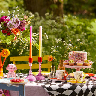 A vibrant outdoor table setting featuring the Alice in Wonderland Bright Tea Cup Set by Talking Tables, complete with flowers, candles, cupcakes, a cake, tea sets adorned with tea cups and saucers on patterned tablecloths, all surrounded by lush greenery.