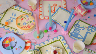 Colorful array of decorated paper plates, cups, and candles with abstract face designs on a pink surface. Various small decorative items and cool Art Party Napkins by Pop! Party Supplies with artistic prints are scattered around a partially eaten cake.