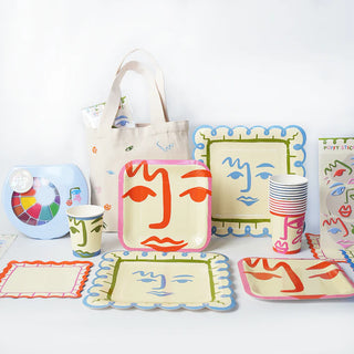 An assortment of tableware featuring abstract face designs, including plates, cups, and Art Party Napkins made from sustainable FSC paper by Pop! Party Supplies, is displayed alongside a tote bag and other accessories adorned with artistic prints, all arranged on a white surface.