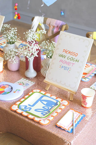 A table is decorated for an art-themed party with plates, cups, and napkins featuring colorful designs. The centerpiece is a canvas on a small easel made by Pop! Party Supplies called the Art Party Paper Pad with the names Picasso, Van Gogh, and Monet, perfect for inspiring budding artists.