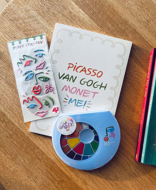 Art supplies on a table include a set of puffy stickers with various shapes, a round watercolor palette, and an Art Party Paper Pad by Pop! Party Supplies with "Picasso, Van Gogh, Monet, ME!" written on the cover. Perfect for budding artists!