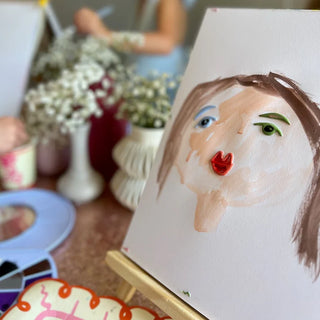 A handmade painting of a face with colorful accents on display, adorned with Pop! Party Supplies Art Party Puffy Stickers in the background alongside flower vases and painting supplies.