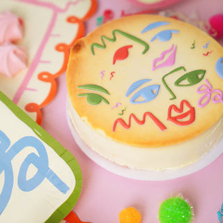 A cheesecake decorated with colorful, abstract face designs sits on a pastel pink table. Nearby are two pink pastries and vibrant decorations, including a paper plate adorned with Pop! Party Supplies Art Party Puffy Stickers and fluffy pom-poms.