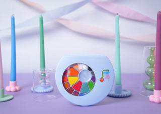 A stunning Pop! Party Supplies Art Party Watercolor Palettes with 24 vibrant hues is artfully displayed on a purple surface, surrounded by pastel-colored candles and elegant glass holders, creating an Art Party atmosphere.