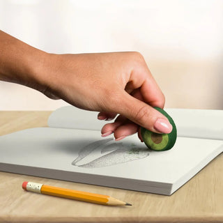 A hand using an avocado pit as an eraser on a sketchbook, perfectly illustrating the innovative Avocado Erasers by Fred and Friends. A pencil lies on the table beside the sketchbook.