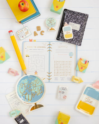 An overhead view of school supplies on a white surface, featuring a My Mind’s Eye Composition Notebook Plate along with a notebook, pencil, erasers, glue, and a planner open to a maze and word search page. An apple sits on other supplies in the top left corner.