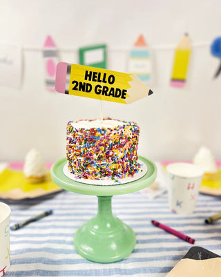 A small sprinkle-covered cake on a green stand, featuring Merrilulu's "Back To School Custom Cake Topper" that reads "Hello 2nd Grade," is surrounded by colorful crayons and paper cups on a striped tablecloth. Perfect for a back-to-school party!