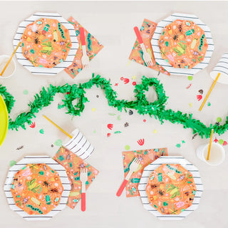 A table is set with small, vibrant Backyard Bugs plates from Daydream Society, along with colorful tropical-themed napkins and cups. Green decorative pieces run through the center like vibrant neon bugs, accompanied by yellow straws paired with the cups.