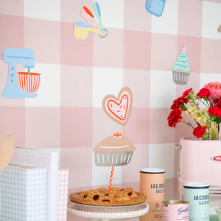 A pie on a table with cupcake decoration evokes holiday cheer, surrounded by the Daydream Society's "Baking Spirits Bright Baking Banner," baking-themed wallpaper, books, a pink toaster, and salt containers. Red flowers in a vase add festive charm to the scene.