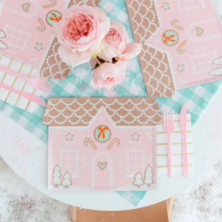 The table is set for a cookie decorating party with Daydream Society's Baking Spirits Bright Pink Gingerbread House Large Plates, pink cutlery, and adorned with roses on a checkered cloth, bringing holiday cheer and baking spirits bright.