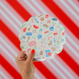 A hand holds a Daydream Society's "Baking Spirits Bright Scalloped Small Plate," featuring baking-themed illustrations, set against a red and white striped background, radiating holiday cheer.