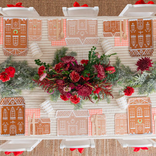 A top view of a festive feast table setting features Baking Spirits Bright Tall Gingerbread House Guest Napkins by Daydream Society, accompanied by red flowers and a greenery centerpiece that exudes holiday cheer. White chairs adorned with red bows complete the enchanting scene.