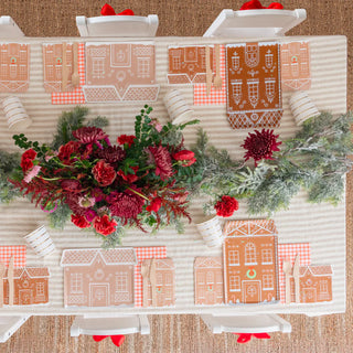 Holiday cheer abounds in this festive table setting, featuring red flowers and a greenery centerpiece. Gingerbread house placemats add charm, while plaid napkins complement the white chairs. It's the perfect scene for a "Baking Spirits Bright" celebration with Daydream Society's Baking Spirits Bright Tall House Large Plates or a cozy cookie decorating party.