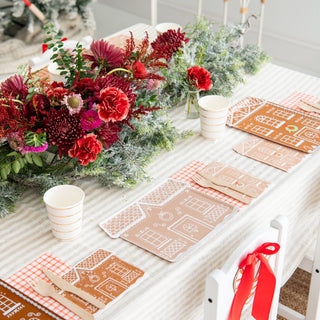 The festive table setting radiates holiday cheer with a red and green floral centerpiece, Daydream Society's Baking Spirits Bright Tall House Large Plates, napkins, and paper cups on a striped tablecloth—ideal for a Baking Spirits Bright cookie decorating party.