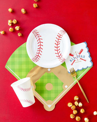 A baseball-themed celebration setup with party essentials including My Mind’s Eye Baseball Paper Plates, a napkin saying "All Star," a white cup with red stitching, a gold fork, and scattered popcorn on a vibrant red background.