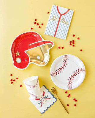 Baseball-themed party essentials including helmet-shaped plates, My Mind’s Eye Baseball Paper Plates, a cup with a napkin, a card, and scattered red candies on a yellow background.