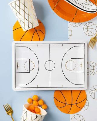 A table decorated with basketball-themed plates, napkins, and My Mind’s Eye Basketball Paper Party Cups. A platter designed like a basketball court is in the center, surrounded by orange napkins, a bowl of cheese puffs, and utensils. Ideal for a child’s party, it even features a charming basketball net design on the cups.