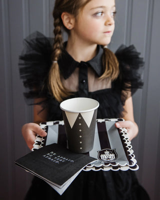 A young girl with braided hair holds a tray featuring a black cup designed as a tuxedo and My Mind’s Eye's Be Afraid Paper Cocktail Napkins. She is dressed in a black outfit and standing against a gray backdrop.