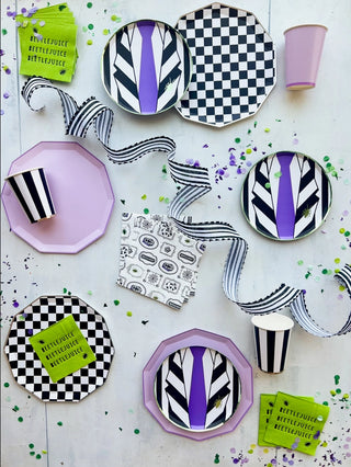 A table set with Beetlejuice-themed party supplies, including plates, cups, eco-conscious Beetlejuice X Bonjour Fête Small Napkins from Bonjour Fête, and ribbon, featuring black-and-white stripes, checkered patterns, and green accents.