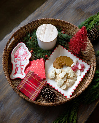 A wicker tray brimming with cookies, a candle, pine cones, and the festive mood of the season. It features a Santa-themed plate, checkered holiday cocktail napkins including My Mind’s Eye Believe Dot Cocktail Napkins and some with scalloped edges, along with a small red Christmas tree figurine.