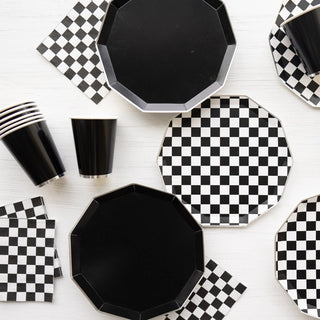 A collection of **Bonjour Fête Black and White Signature Checker Dinner Plates**, checkered napkins, and black cups are meticulously arranged on a light surface.