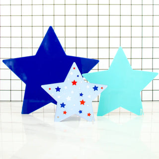 Three Blue and Turquoise Acrylic Stars Decor by Kailo Chic of varying sizes and colors (blue, light blue, and white with red and blue stars) are displayed against a white grid background. Perfect for 4th of July celebration, these star-shaped objects add a festive touch to your home decor.