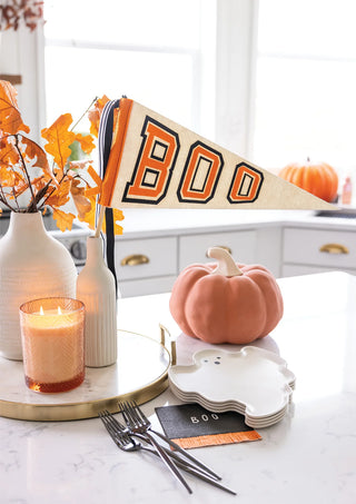 Festive Halloween decor featuring My Mind’s Eye "Boo Felt Pennant Banner," fall leaves in vases, a lit candle, a pink pumpkin, ghost-shaped plates, and silverware on the counter—perfect for any Halloween celebration.