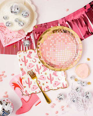 Overhead view of a festive table setting with My Mind's Eye cowgirl pattern paper plates, a red napkin, disco balls, and decorative confetti on a white surface.