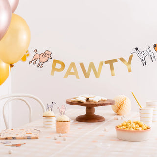 A festive "pawty" setup with Daydream Society Bow Wow Thingamajigs garland decoration featuring cute dog illustrations, surrounded by balloons, snacks on a wooden stand, and party cups, all set against a soft pink.