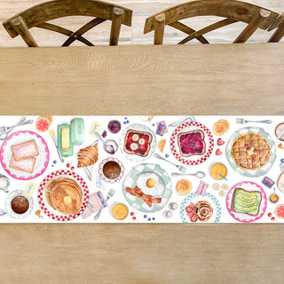 A wooden table is adorned with the vibrant "Breakfast At the Diner Brunch Table Runner" by Cami Monet, showcasing a delightful array of breakfast foods such as waffles, pancakes, bacon, eggs, fruit, and drinks. Four wooden chairs are visible around the table.