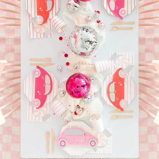 A table set for Valentine's Day with car-themed pink and red decorations includes Buggy of Love Small Plates from Daydream Society, complemented by disco balls, striped straws, and wooden cutlery on a checkered tablecloth.