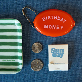 A charming vintage-inspired accessory display features a striped tray, two coins, an orange oval Coin Pouch labeled "Birthday Money" from Three Potato Four, and a matchbook with the text "Sunday," all arranged on a blue surface.