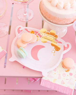 A pink party table featuring a moon-themed cauldron-shaped plate from My Mind’s Eye, holding pink and teal macarons, spooky snacks, patterned pink napkins, and a cake topped with piped frosting.