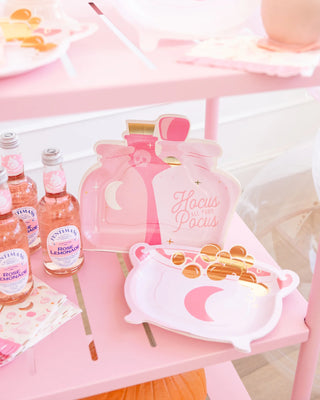 A pink-themed party setup featuring "Hocus Pocus" Halloween party plates shaped like potion bottles, My Mind’s Eye Cauldron Shaped Plates for your spooky snacks, bottles of rose lemonade, and paper cups adorned with moons and stars.