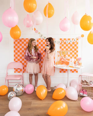 Two girls in party dresses stand among pink and orange balloons, with a checkered backdrop and a banner reading "SPOOKY ERA." A table with snacks, Halloween Checkered Party Cups from My Mind's Eye, and decor is to the right, while balloons scatter across the floor.