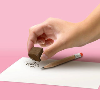 A hand holding a Fred and Friends Chocolate Erasers effortlessly erases pencil marks from a paper placed on a pink surface with a pencil nearby.