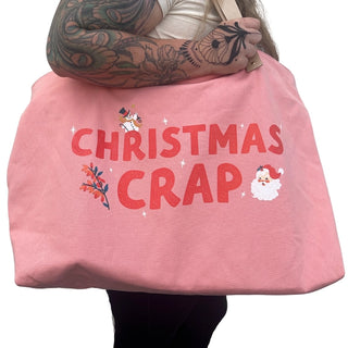 A tattooed person is holding a Fun Club Christmas Crap Oversized Tote Bag, which prominently features the text "CHRISTMAS CRAP" alongside festive illustrations of a snowman, Santa face, and holly leaves.