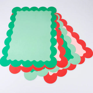 A festive arrangement of Meri Meri's Christmas Paper Placemats in classic red and green scalloped edges, accompanied by pink duo tone paper sheets, rests on a white background.