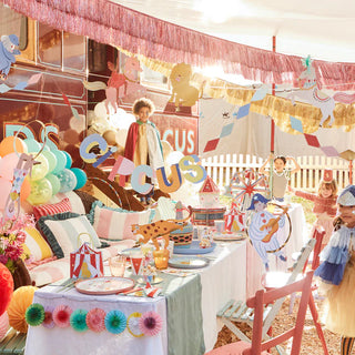 A vibrant Meri Meri circus-themed outdoor children's party with joyous decorations, colorful table settings with gold foil embellishments, and a cheerful child standing by a "circus" sign.