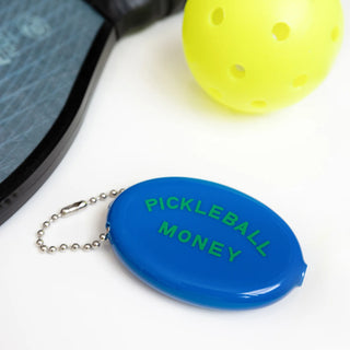 A "Pickleball Money" coin pouch by Three Potato Four, featuring a vintage-inspired blue design, lies next to a yellow perforated pickleball and a pickleball paddle on a white surface.