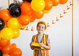 A child dressed as a construction worker, wearing an orange and yellow safety vest and helmet, holds a toy tool. Orange, yellow, and black balloons and mini traffic cones decorate the background at this lively construction-themed event featuring the Construction Cone Banner by My Mind’s Eye.