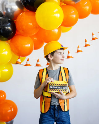 A child in a construction-themed outfit holds a "Caution Treat Zone" sign, surrounded by orange, yellow, and black balloons shaped into an arch. Small orange and white traffic cones complement the festive My Mind’s Eye Construction Cone Banner in the background.