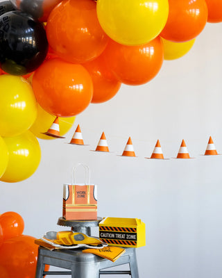 A party setup features a vibrant My Mind's Eye Construction Cone Banner with yellow, orange, and black balloons, and miniature traffic cones strung as bunting. A small gift bag labeled "Work Zone" and a "Caution Treat Zone" box complete this construction-themed event decor on a stool.