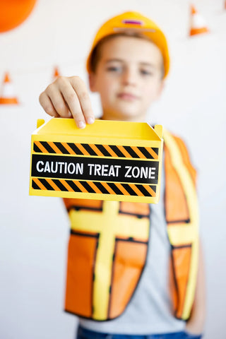 A young person wearing a construction hat and safety vest holds a yellow box labeled "Construction Toolbox Treat Containers" by My Mind’s Eye, perfect for a construction-themed party.