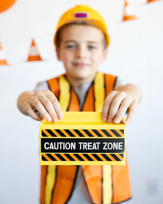 A child wearing a construction worker costume holds a yellow and black box labeled "Construction Toolbox Treat Containers" by My Mind's Eye, making it the perfect party favor for a fun-filled construction party.