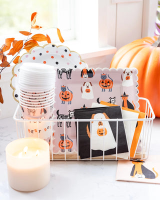 A white wire basket holds Halloween-themed plates and My Mind's Eye Halloween Pet Costume Cocktail Napkins, featuring ghost and pumpkin designs, next to a stack of plastic cups, a lit candle, and a decorative pumpkin on the table.