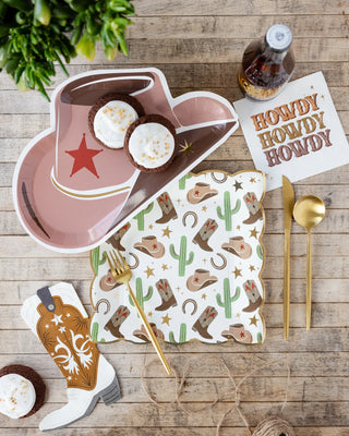 Western-themed table setting featuring cowboy hat and boot-shaped plates, My Mind’s Eye Cowboy Boot Shaped Guest Napkin with "Howdy Howdy Howdy" text, a gold fork and spoon, and decorations with cacti and horseshoes—perfect for your Wild West party.