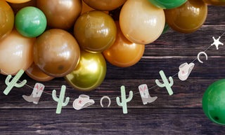 A **Cowboy Icon Banner** by **My Mind’s Eye** hangs below a bunch of green, brown, and gold balloons against a wooden background.